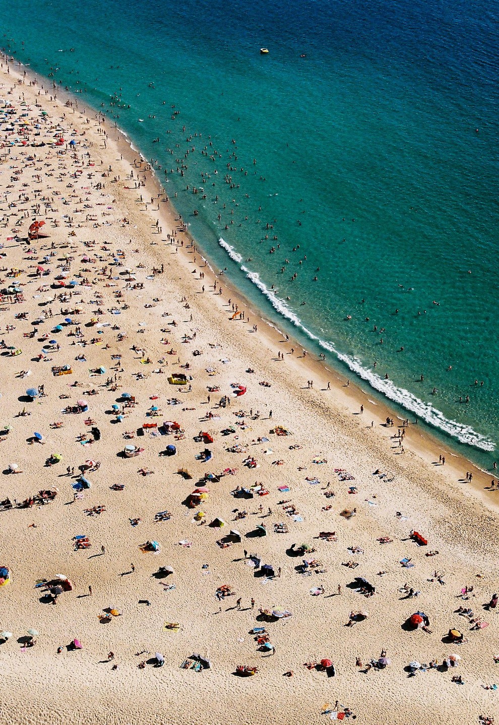 An aerial view shows a crowded beach next to a turquoise ocean with many people swimming.