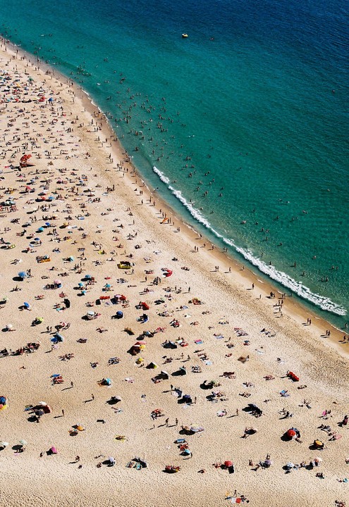 An aerial view shows a crowded beach next to a turquoise ocean with many people swimming.