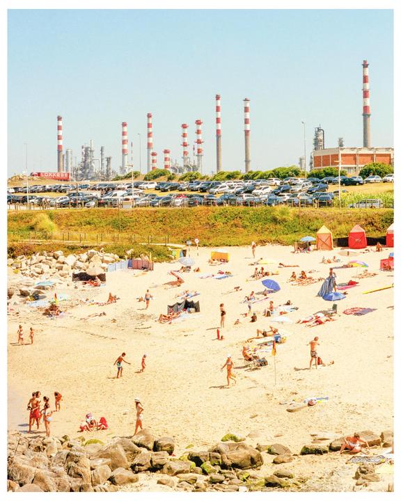 A beach with many people and umbrellas is in the foreground, with a parking lot full of cars and an industrial complex with tall striped smokestacks in the background.