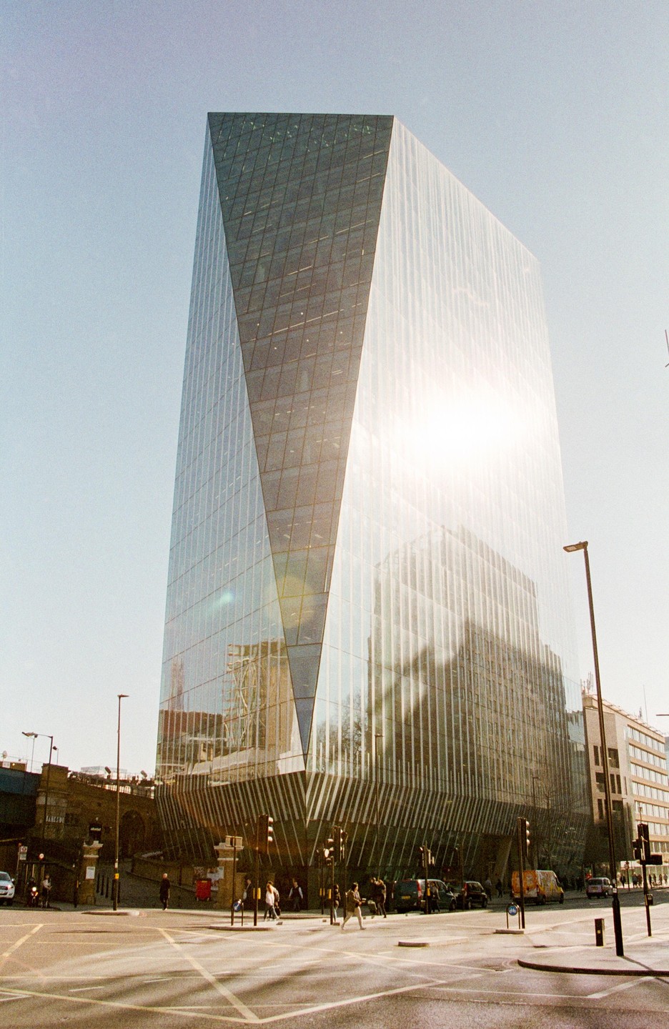 A tall, modern glass building with a unique geometric design stands at a street intersection under a bright sky.