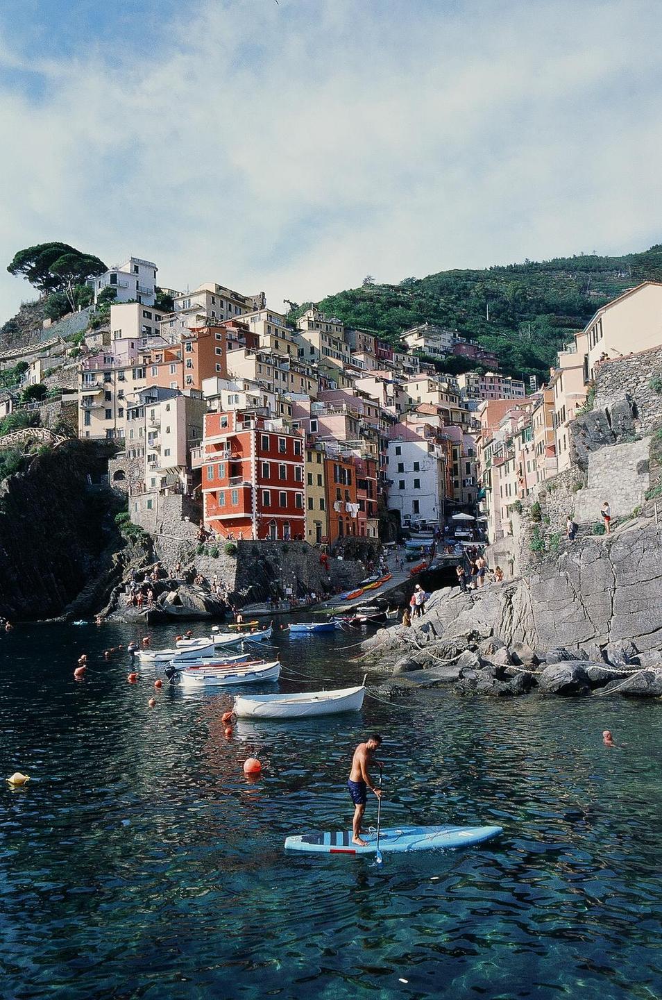 A man paddleboards in a harbor with colorful buildings on a hillside in the background.