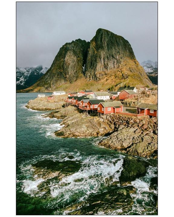 Red and white houses on stilts line a rocky coast with a large mountain in the background.