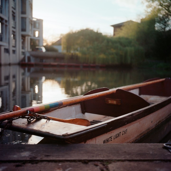 A wooden boat with oars rests on a canal with buildings and trees in the background.