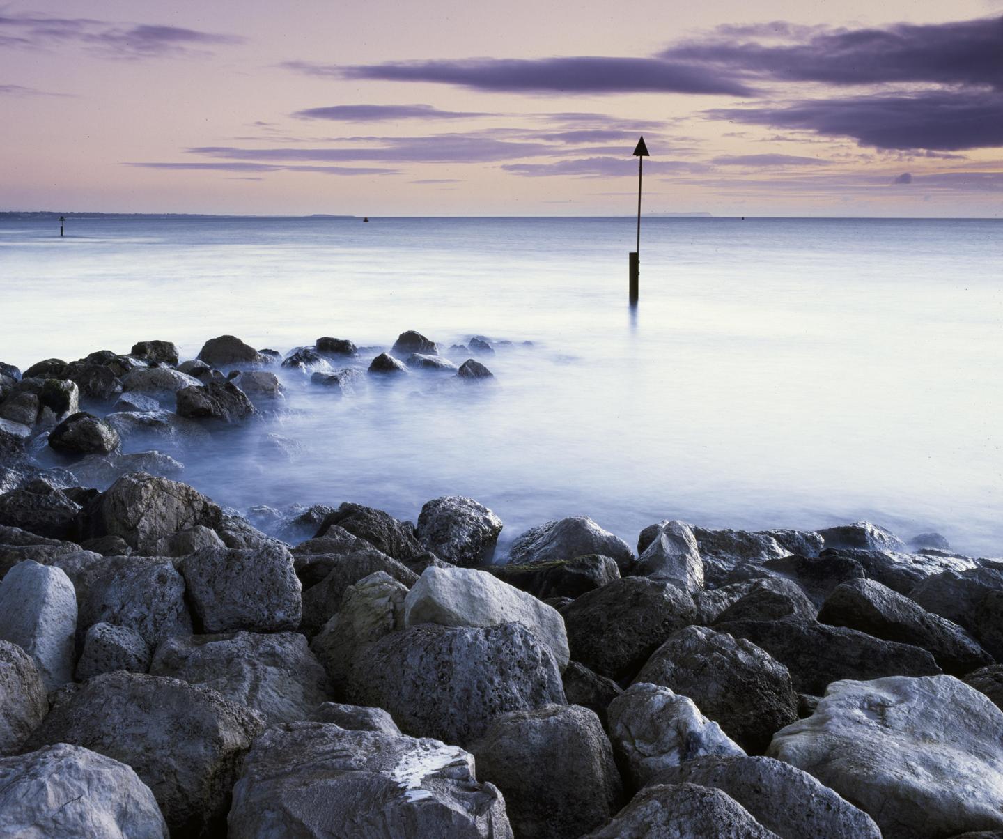A rocky shore meets a calm sea under a sky with purple clouds.