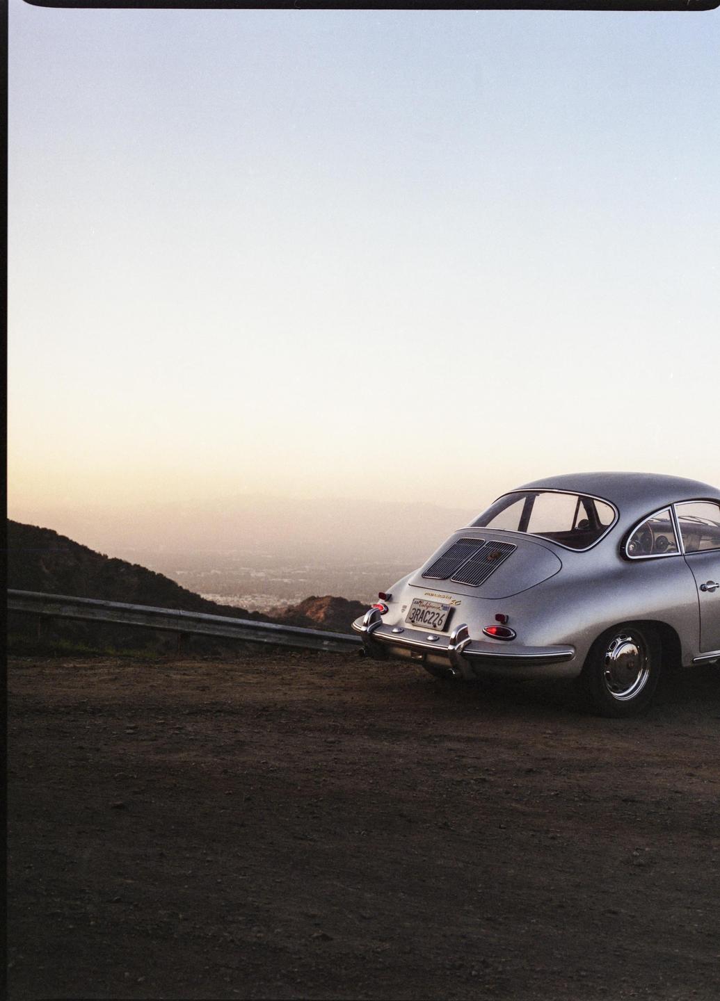 A silver car is parked on a dirt road overlooking a city at dusk.