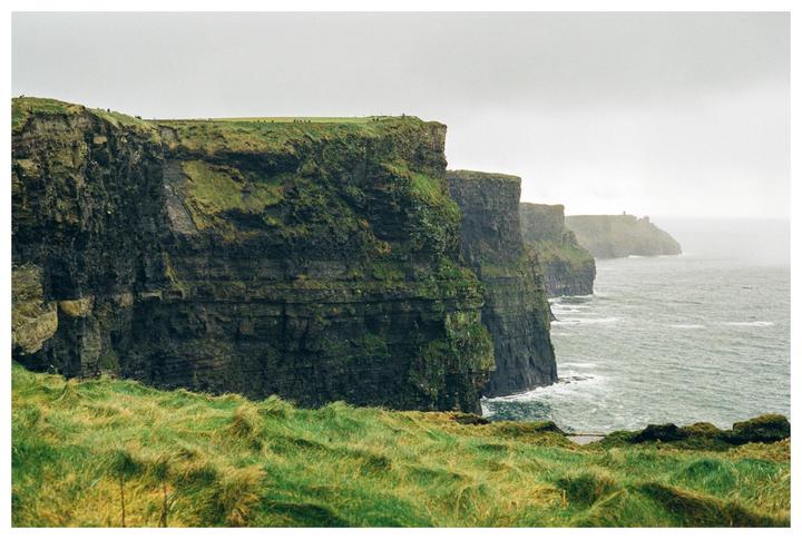 A dramatic landscape shows the Cliffs of Moher with green grass in the foreground and a hazy ocean.