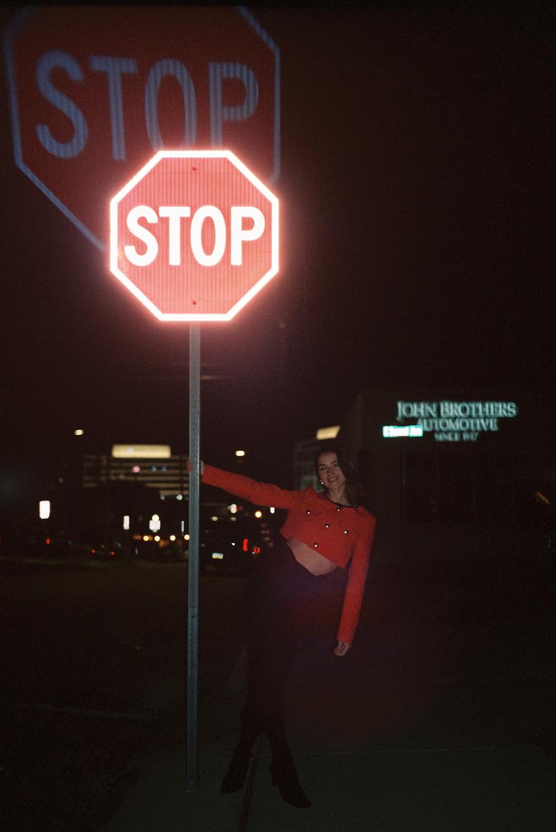A woman in a red top stands next to a stop sign at night, with a faint reflection of the sign visible above it.