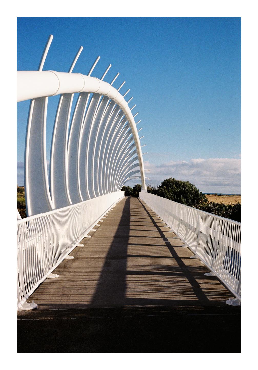 A white pedestrian bridge with an arched, ribbed structure casts long shadows on the walkway under a blue sky.