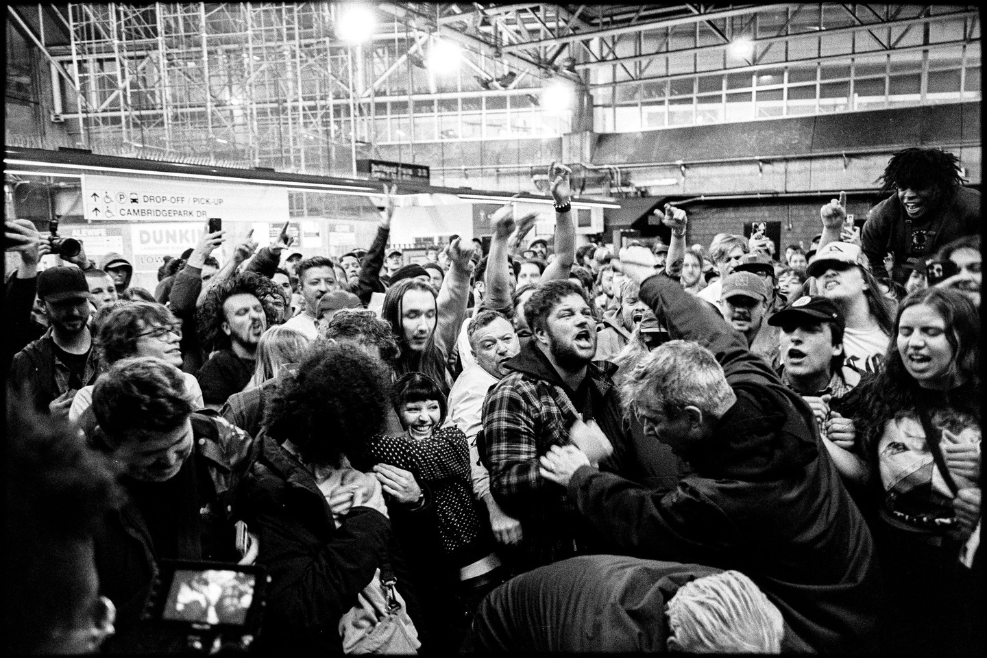 A large crowd of people are gathered in what appears to be a train station, with many raising their hands and some looking excited or singing.