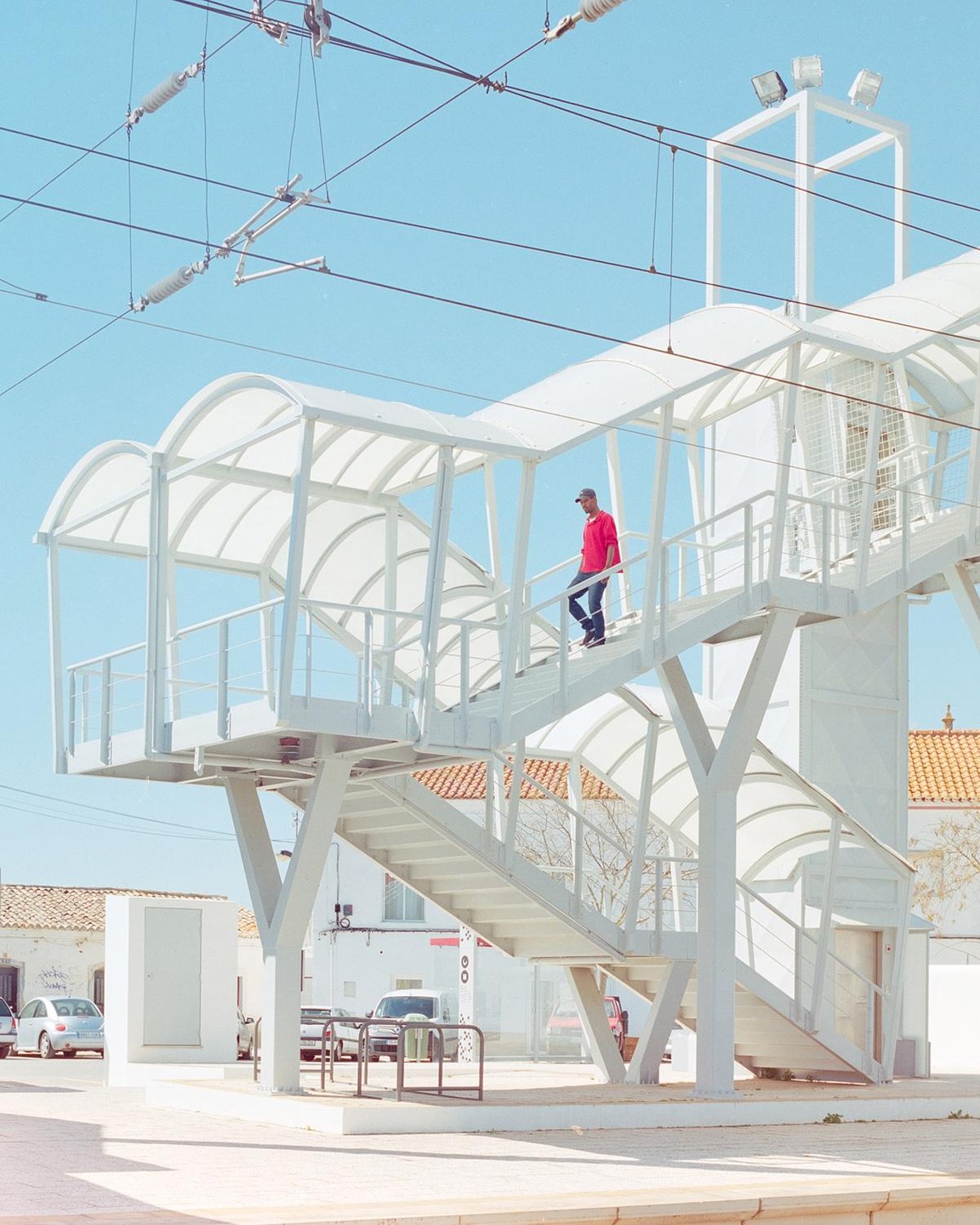 A man in a red shirt walks down a white pedestrian bridge over a street with cars and buildings.