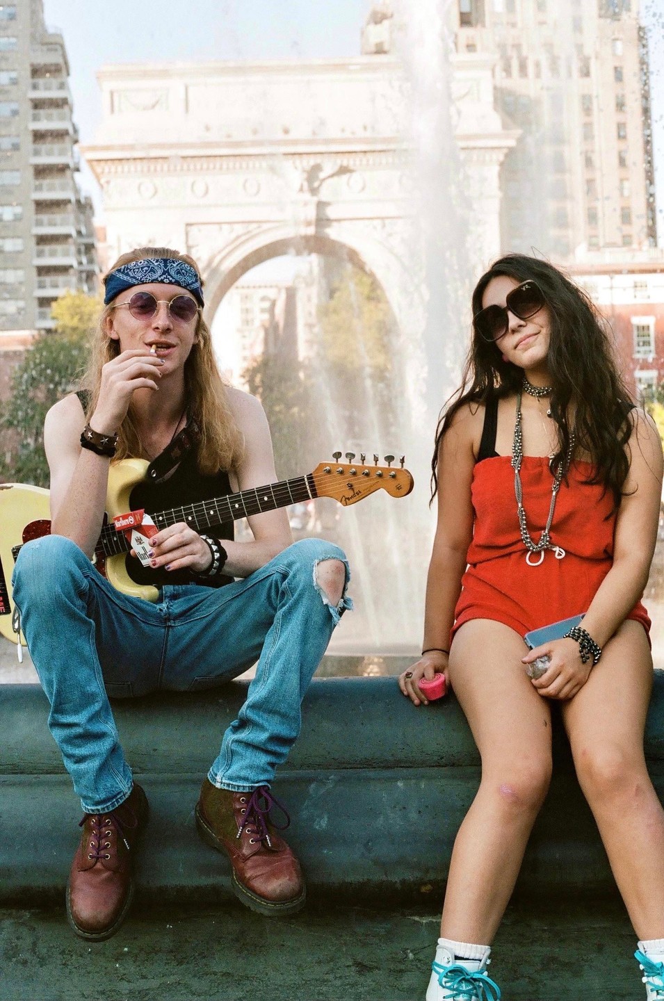 A man with long hair and a woman sit in front of a fountain and an arch in a city park.