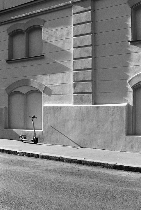 An electric scooter is parked on a sidewalk next to a building with arched windows, casting a long shadow.