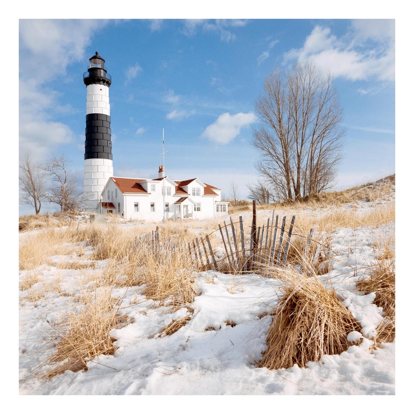 A black and white lighthouse stands next to a white house with a red roof on a snowy hill with dry grass and a wooden fence.