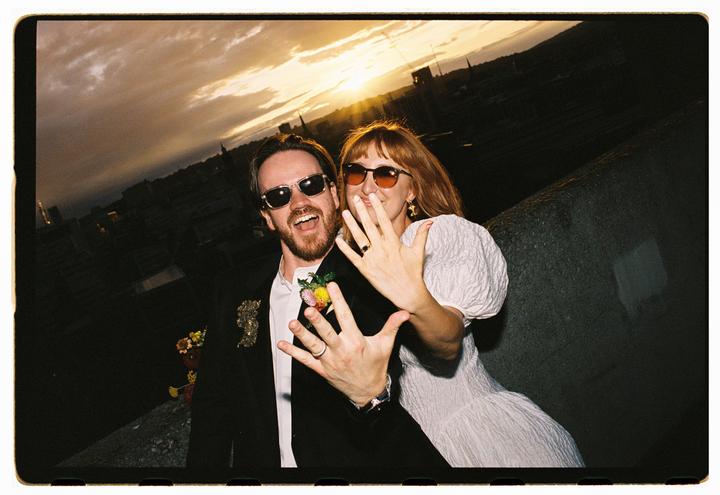 A couple wearing sunglasses shows off their rings at sunset with a city in the background.