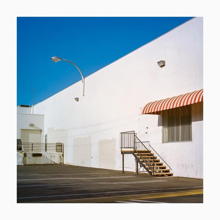 A white building with a red and white striped awning and an external staircase stands next to an empty parking lot under a clear blue sky.