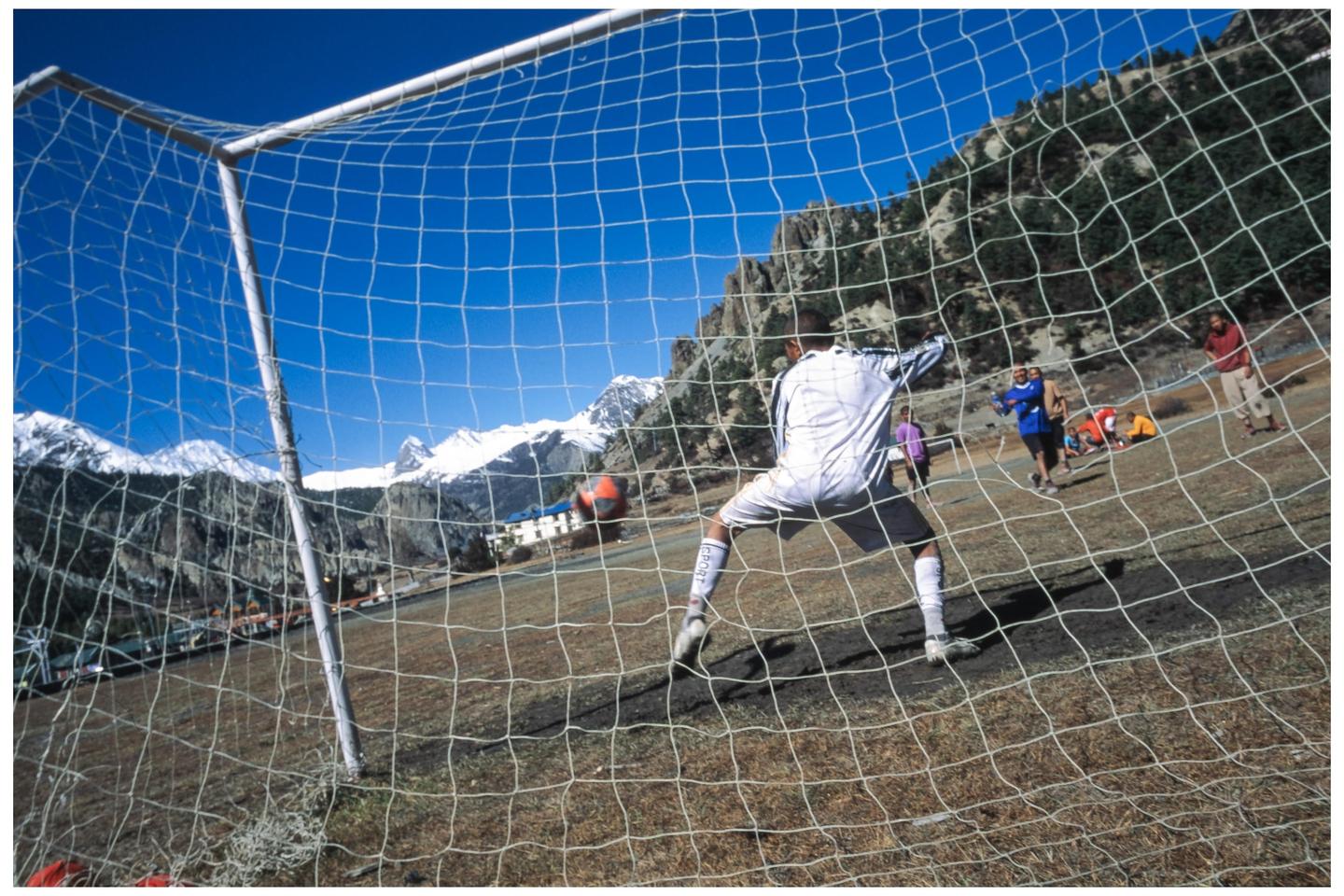 A soccer player in a white uniform is seen through a goal net, playing on a field with snow-capped mountains in the background.