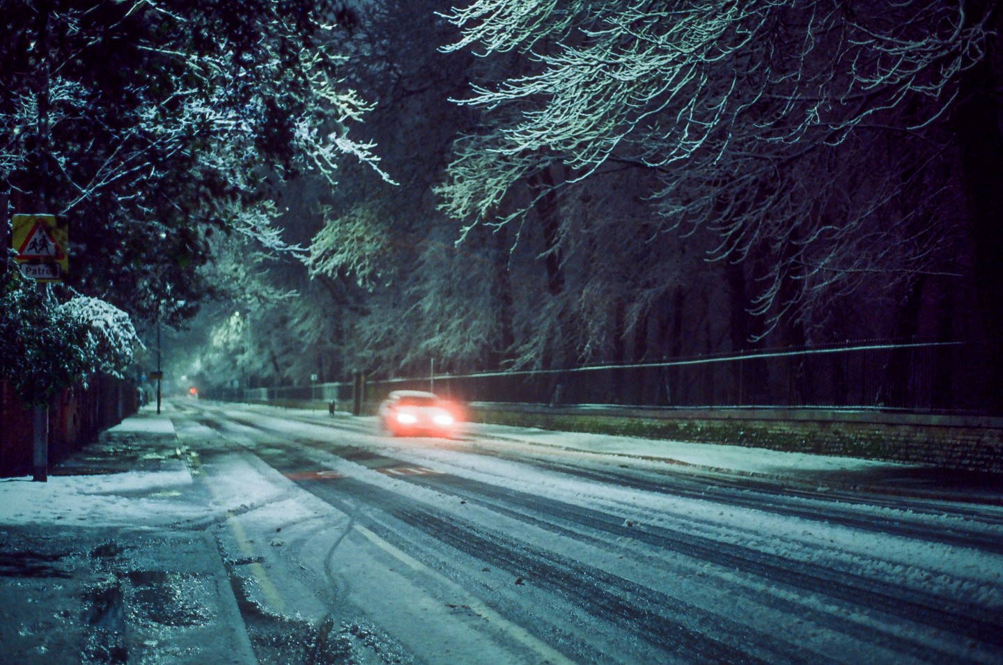 A car with bright red taillights drives down a snow-covered street lined with trees at night.