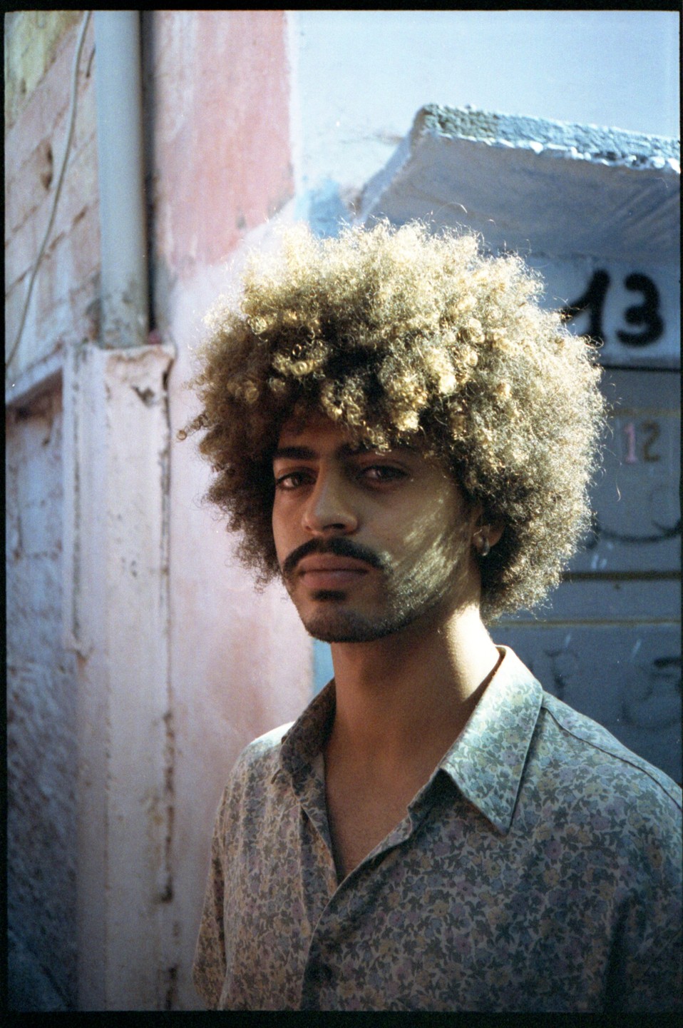 A man with curly hair and a mustache looks directly at the viewer, with a light-colored wall and the number 13 in the background.