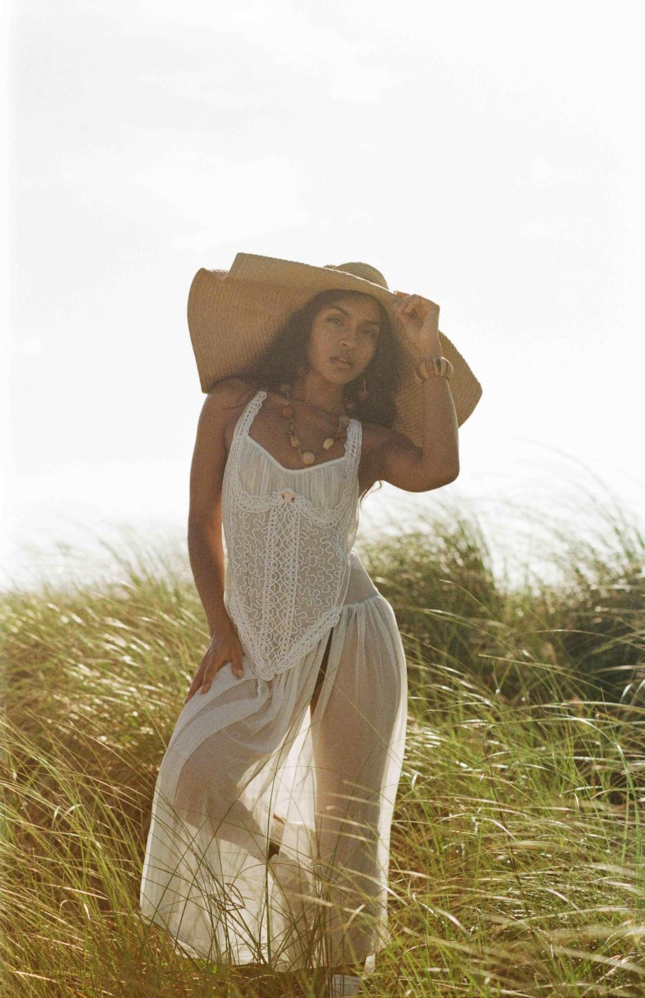 A woman wearing a large straw hat and a white dress stands in tall grass.