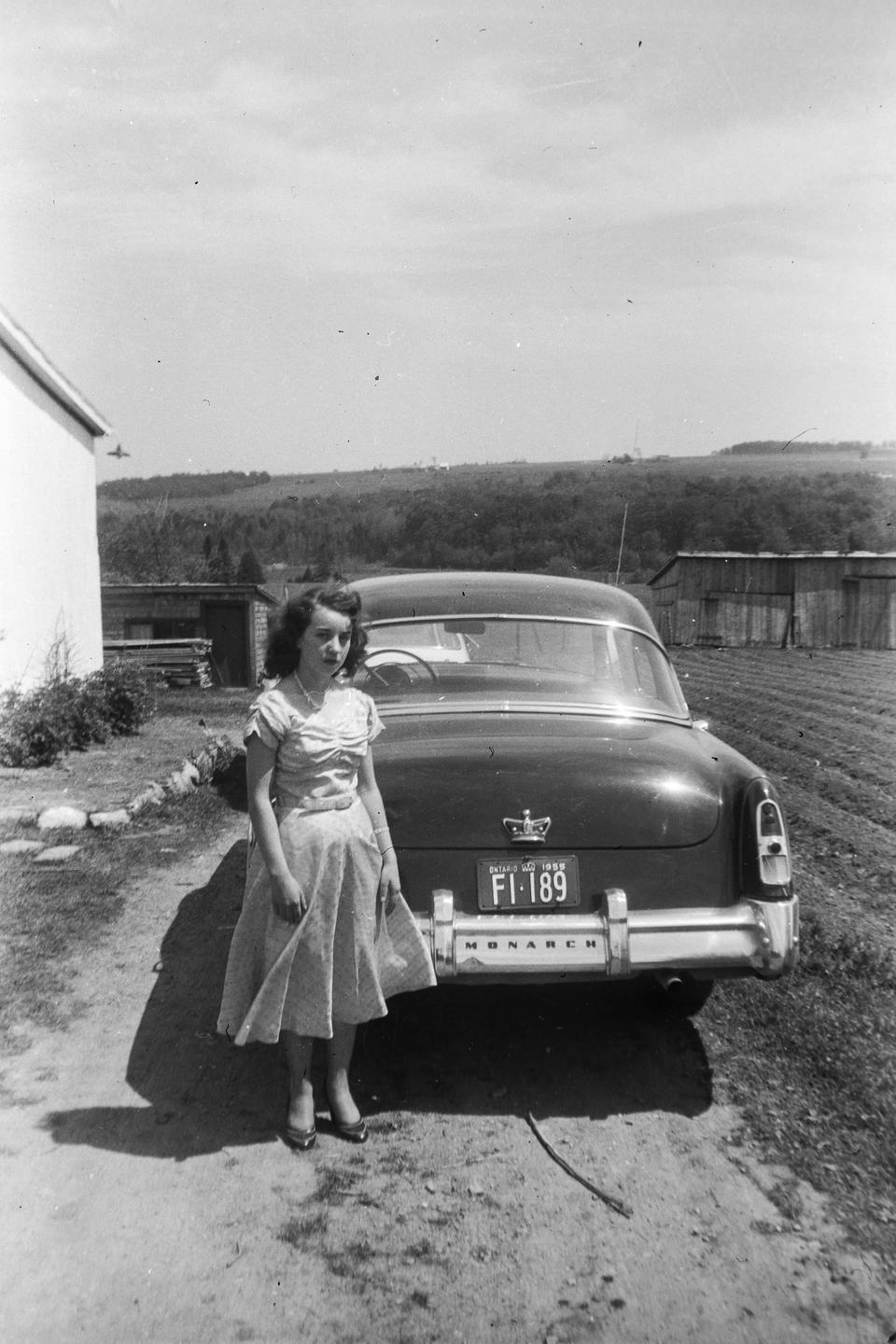 A woman in a dress stands next to a car on a dirt road with a field and hills in the background.