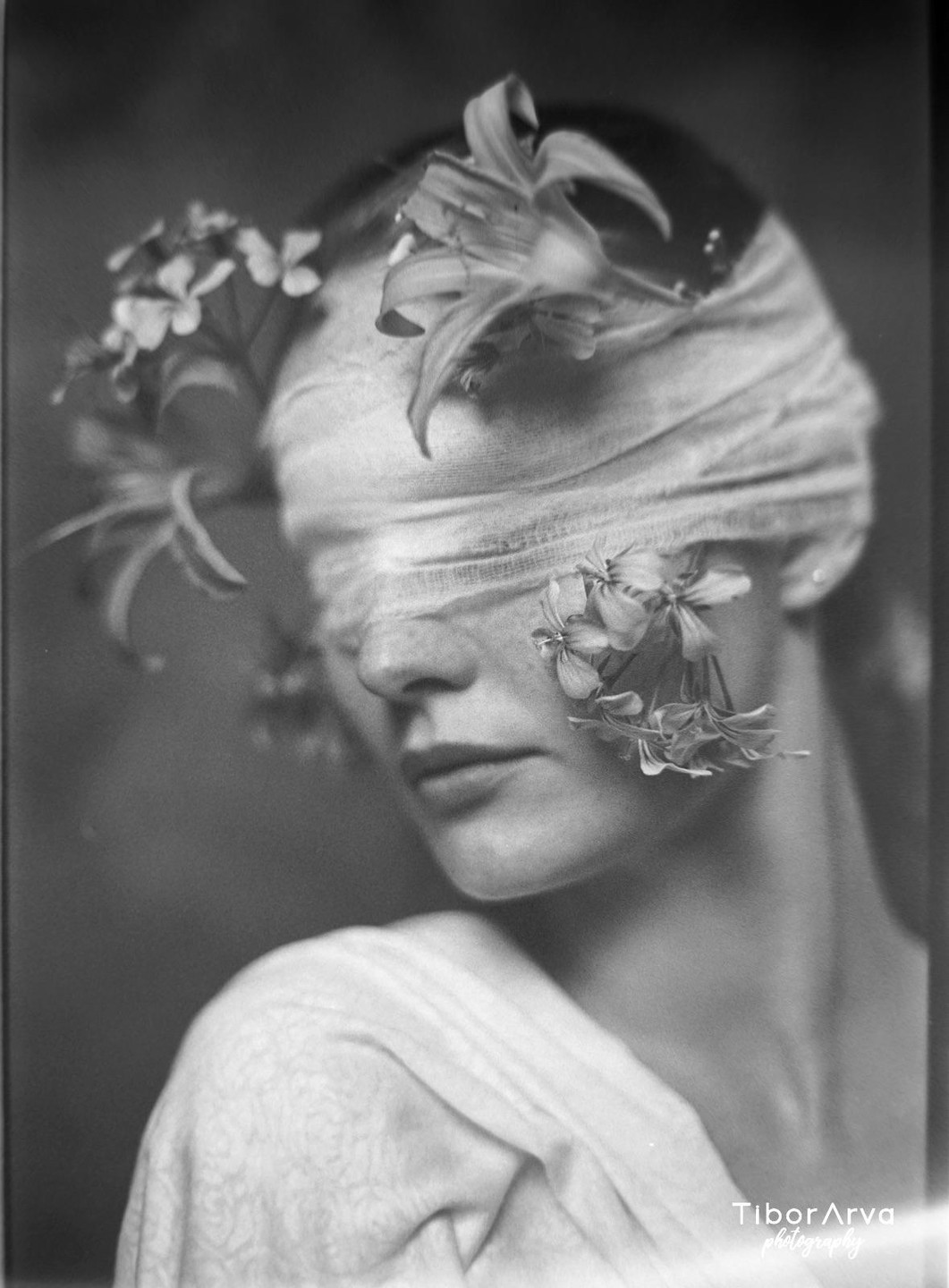 A woman with a blindfold and flowers on her face looks down in a black and white portrait.