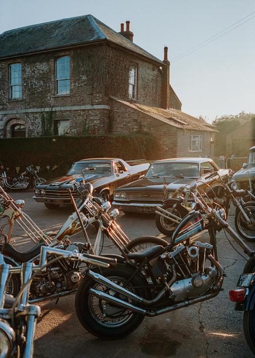 A collection of motorcycles and classic cars are parked in front of a brick building covered in ivy.