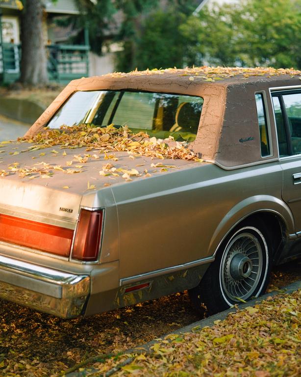 A gold car with a flat tire is parked on a street covered in autumn leaves.