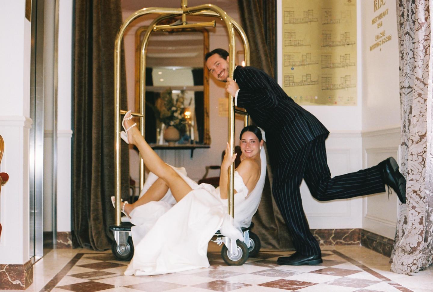 A man pushes a woman in a wedding dress on a luggage cart in a hotel lobby.