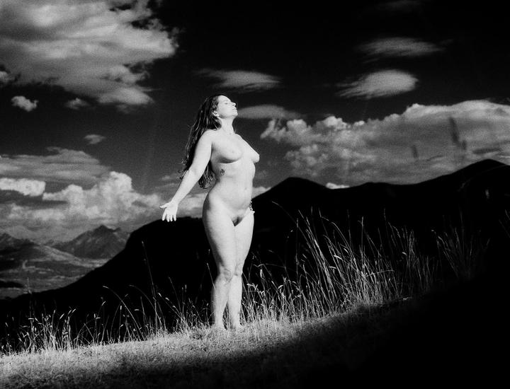 A nude woman stands on a grassy hill with mountains and clouds in the background.