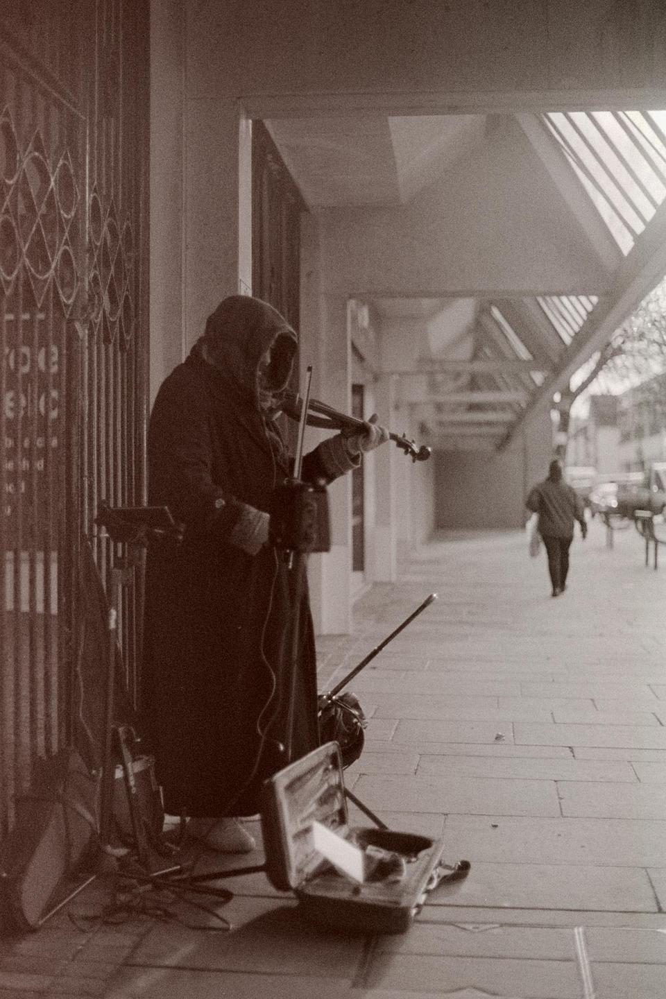 A hooded street musician plays a violin on a sidewalk next to an open violin case.