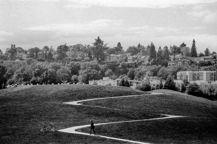 A person walks up a winding path on a grassy hill with houses and trees in the background.