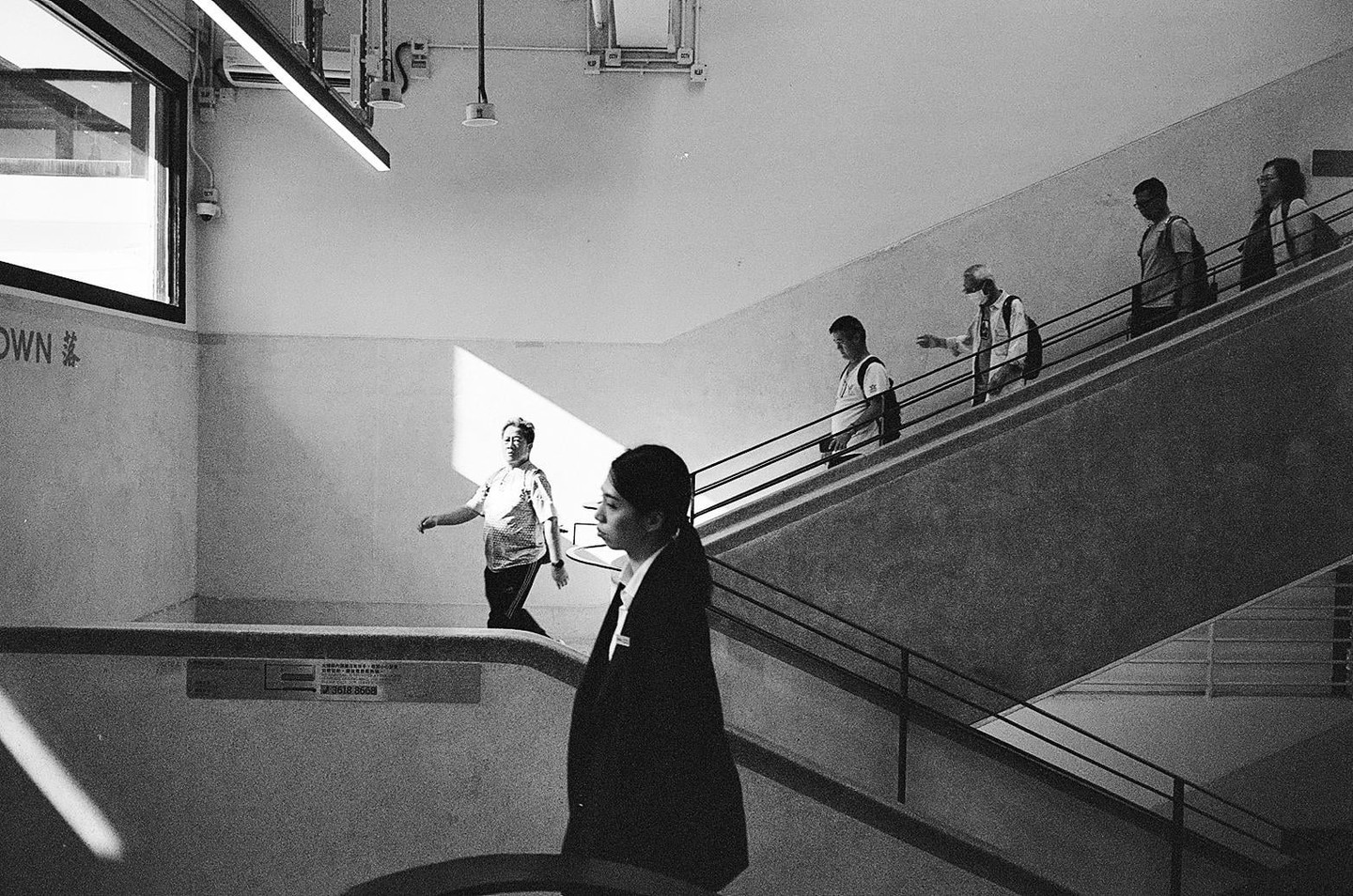 A woman stands in the foreground while other people walk on an escalator and stairs in a building with strong diagonal shadows.