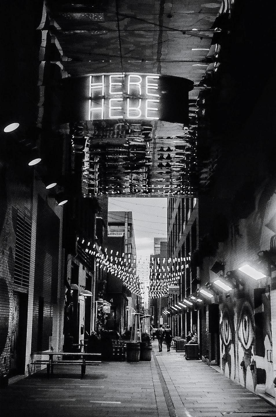 A neon sign reading "HERE HEBE" hangs above a narrow city street decorated with string lights and graffiti.