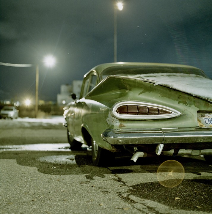 A green vintage car with snow on its trunk is parked on an asphalt surface at night, illuminated by streetlights.