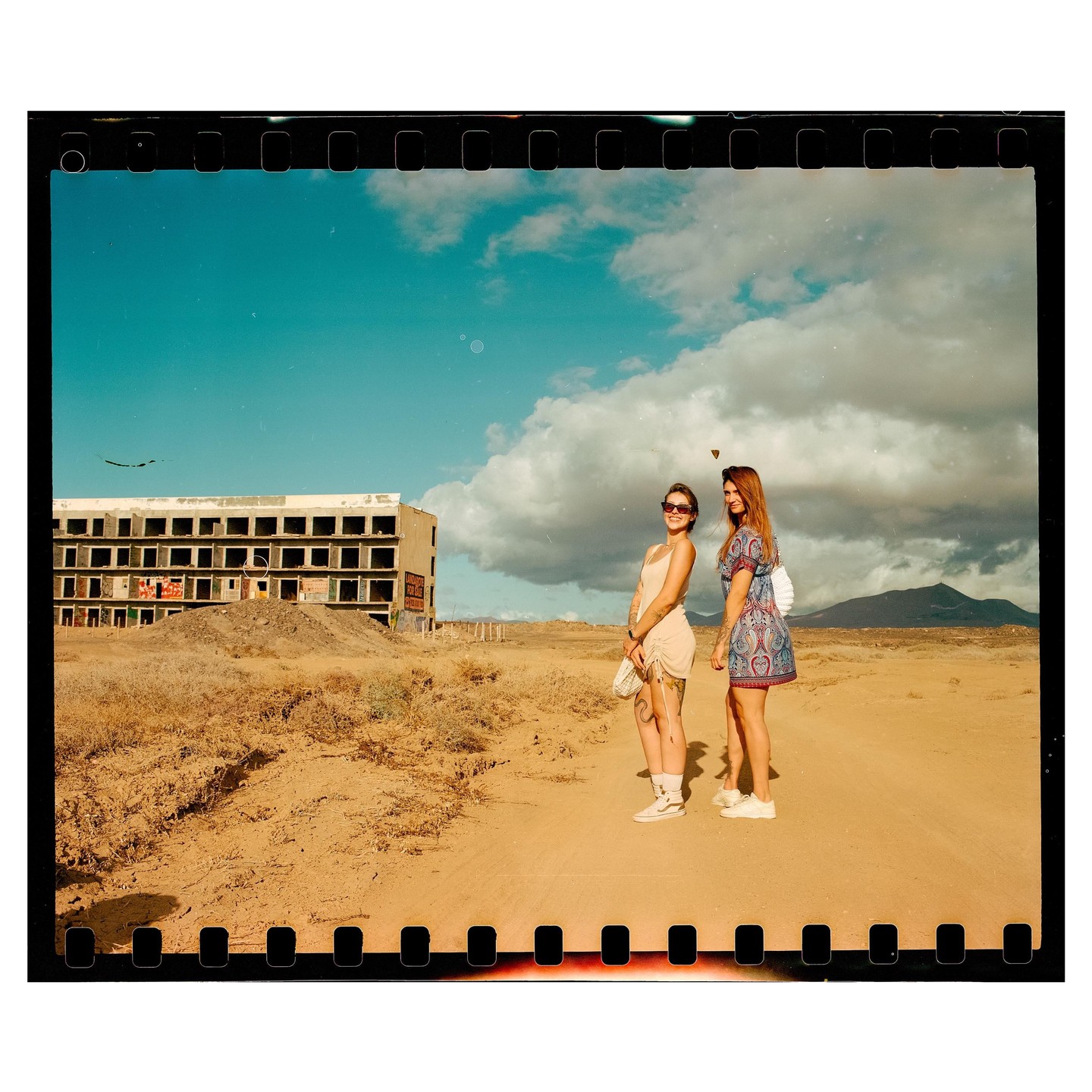 Two women stand on a dirt road in front of an abandoned building under a partly cloudy sky.