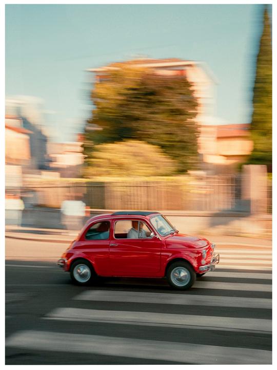A red car with a man driving is captured in motion on a street with a blurred background.