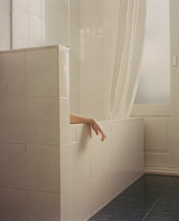 An arm and hand are visible over the edge of a bathtub in a tiled bathroom.