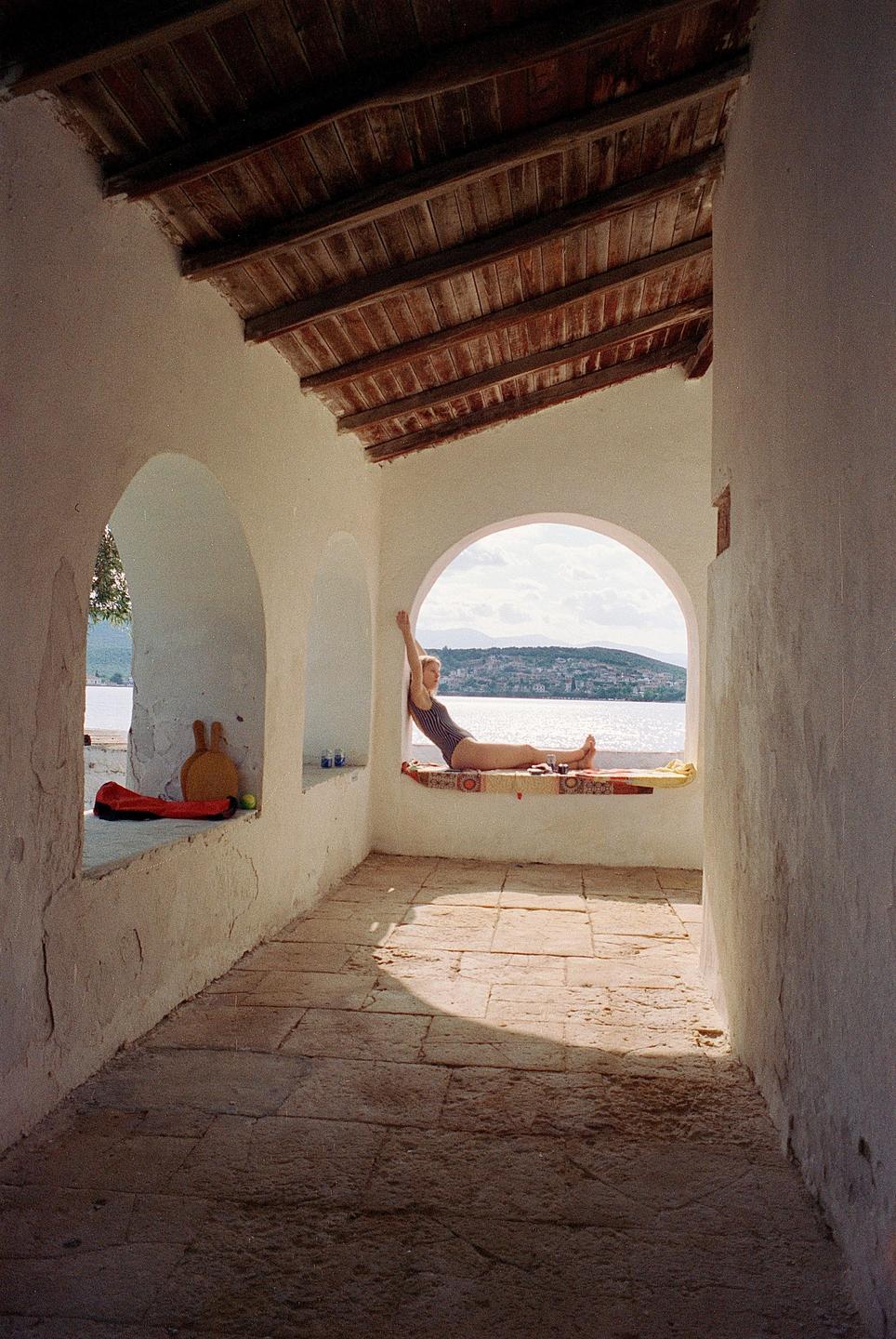 A woman in a swimsuit reclines in an arched window overlooking a body of water and distant hills.