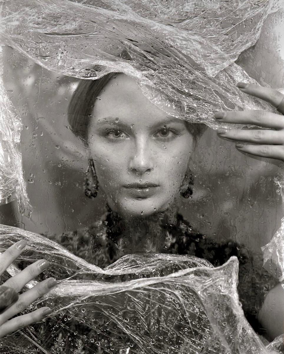 A woman with dark earrings looks directly forward through a plastic sheet covered in water droplets.