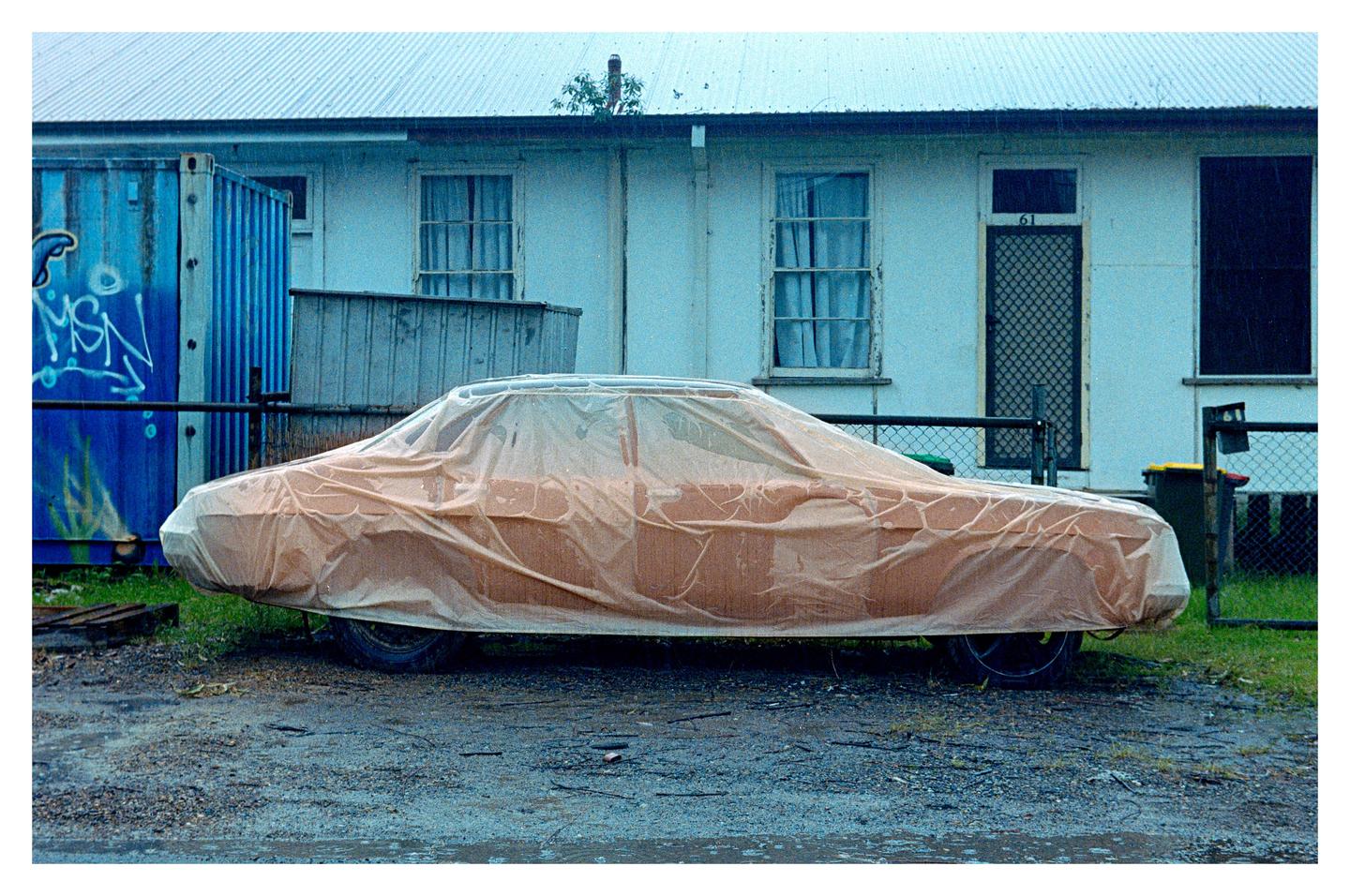 A car covered with a tarp is parked on a dirt patch in front of a white building with a blue shipping container to the left.