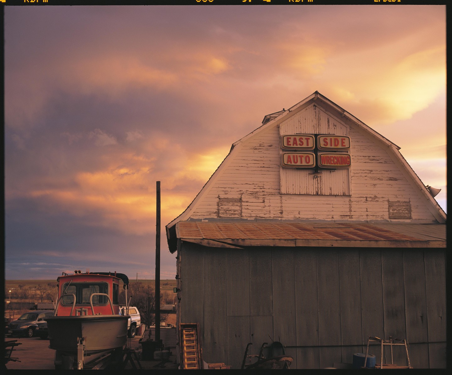 A white wooden building with a red and white sign for "East Side Auto Wrecking" stands next to a boat on a trailer under a dramatic sunset sky.