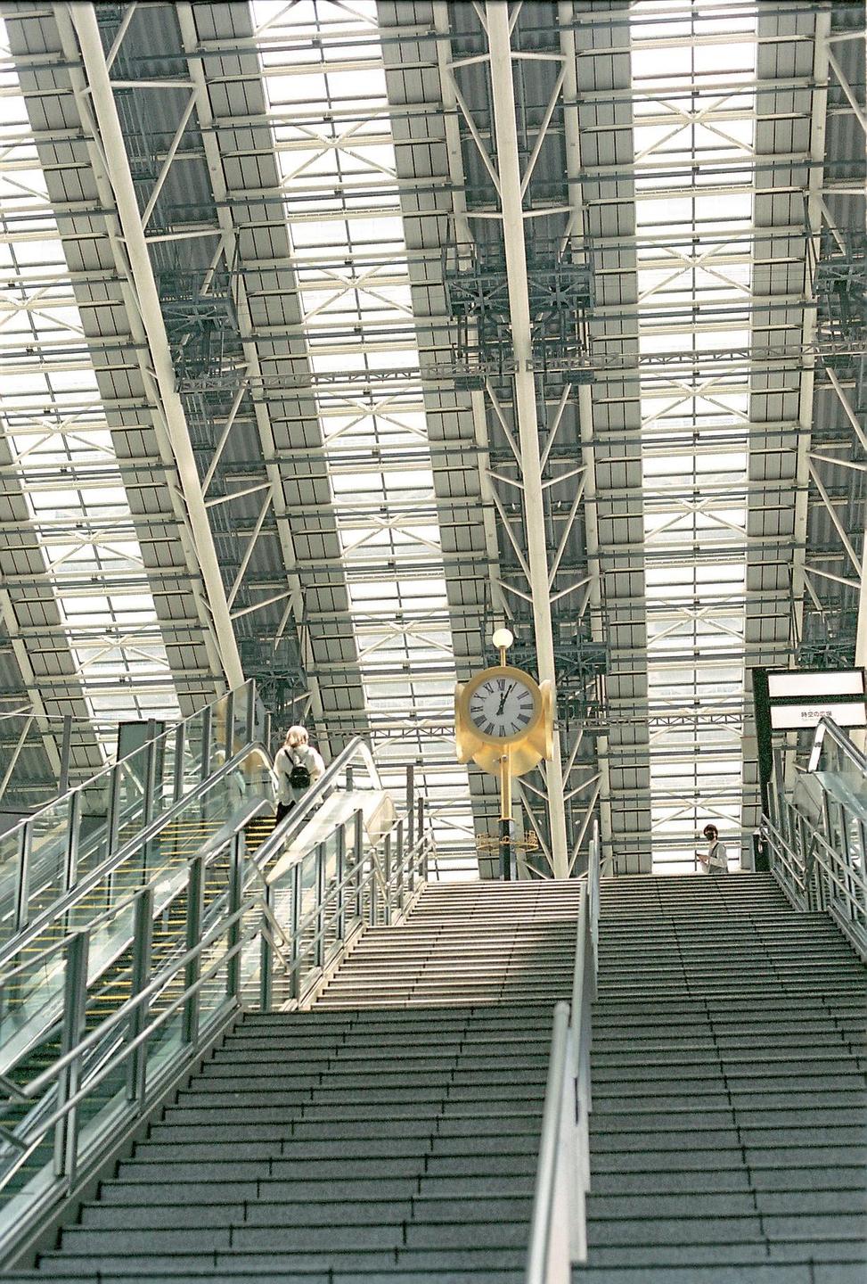 A large golden clock hangs above a wide staircase and escalator in a modern building with a glass and metal roof.