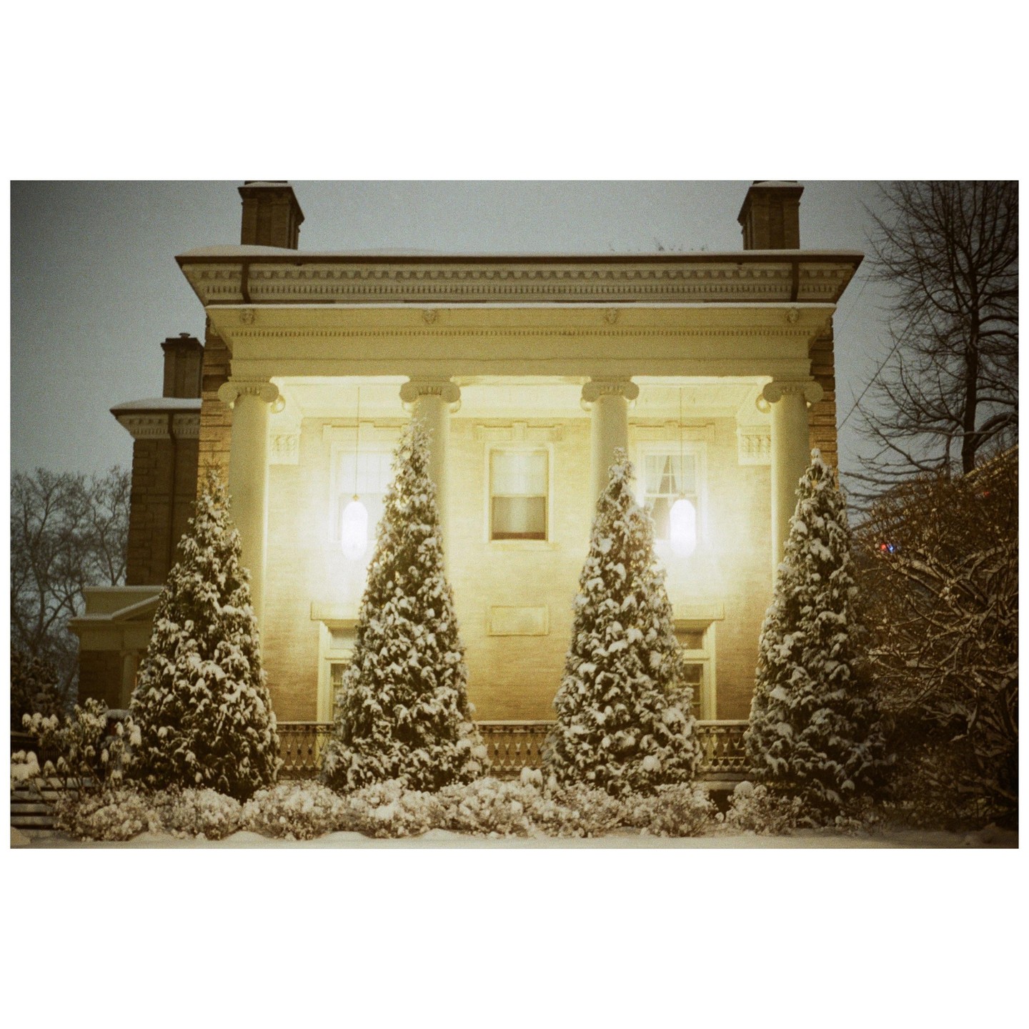A large house with columns and a porch is illuminated at night, with snow-covered evergreen trees in front.