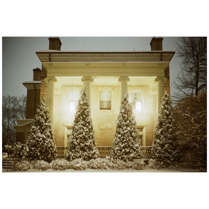 A large house with columns and a porch is illuminated at night, with snow-covered evergreen trees in front.
