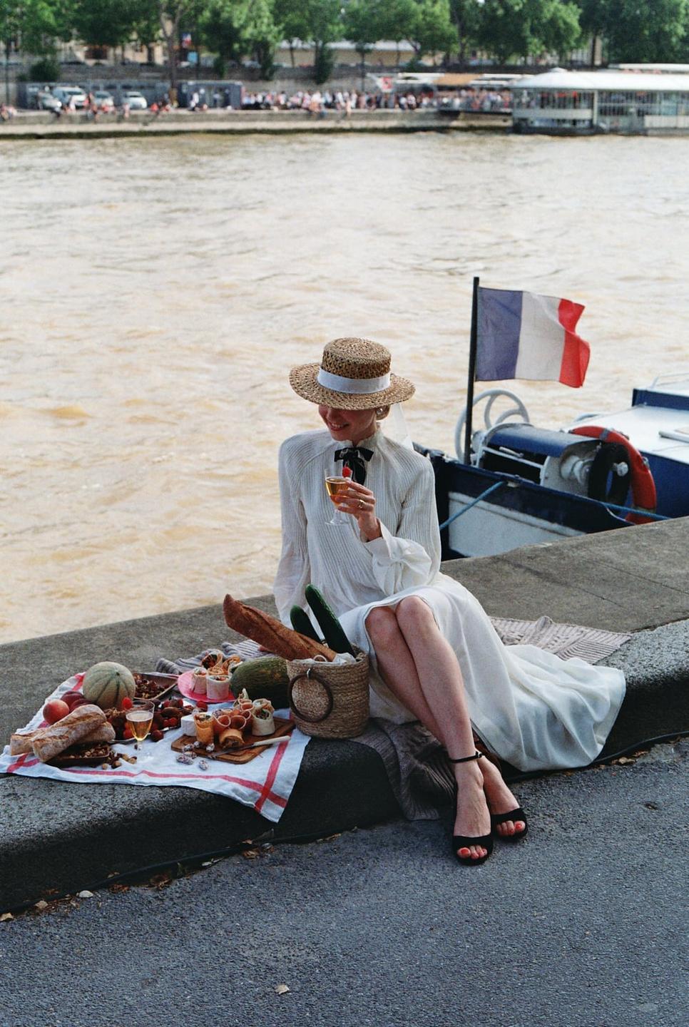 A woman in a white dress and straw hat sits by a river with a picnic spread, a French flag visible on a boat nearby.