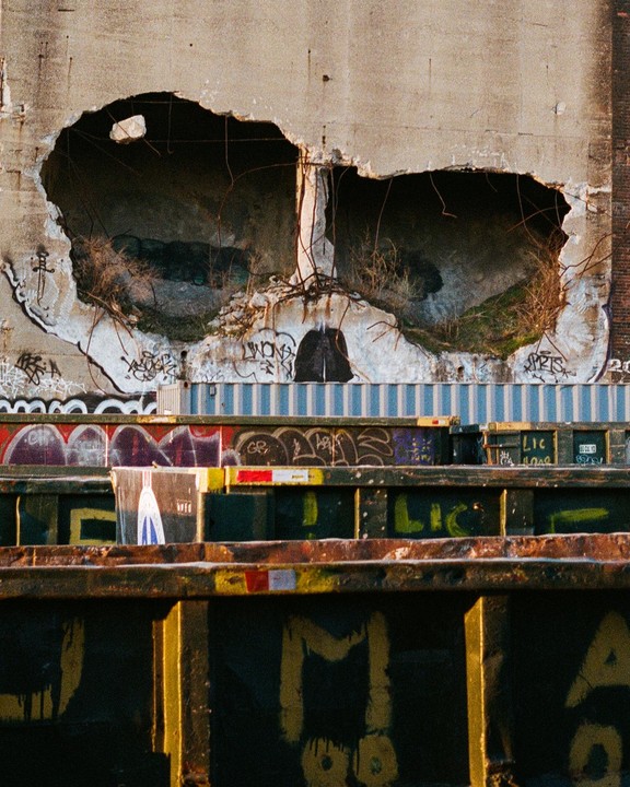 A concrete wall with two large holes resembling eyes is covered in graffiti above several metal containers.