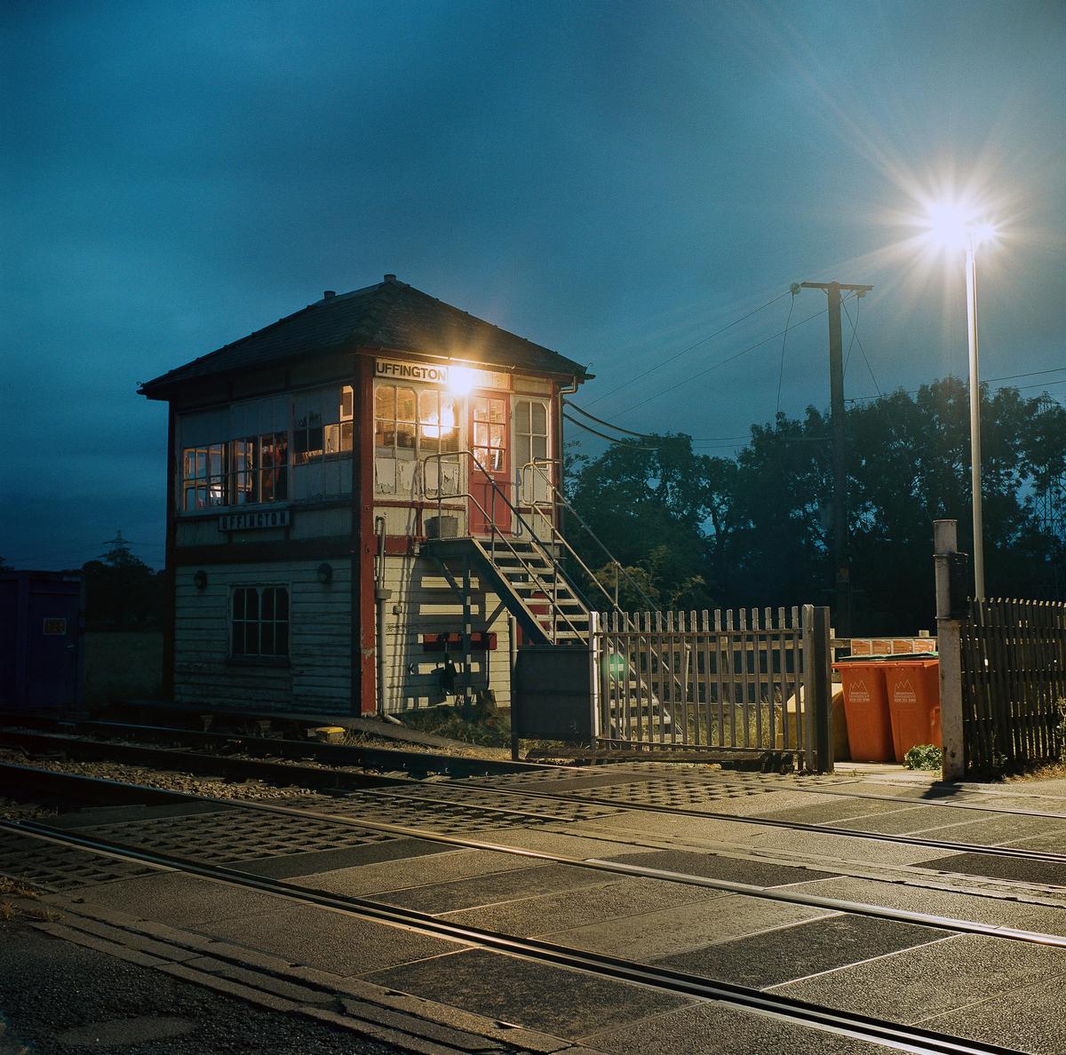 A signal box with illuminated windows stands beside railroad tracks at night, with a bright streetlamp on the right.