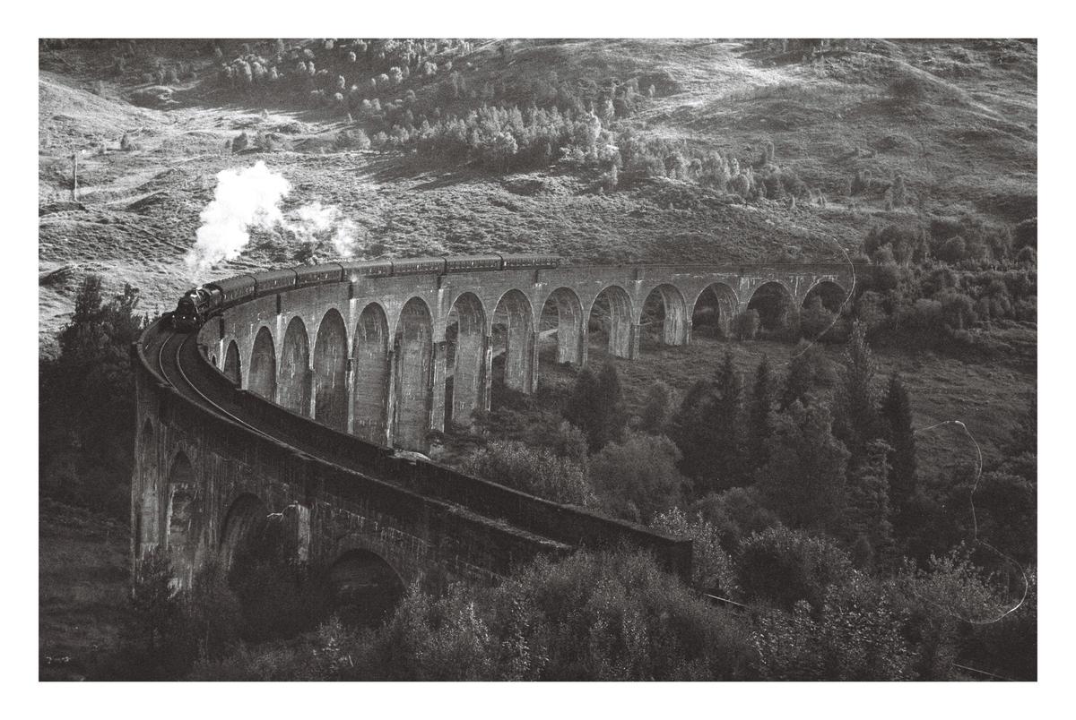 A train with a plume of smoke travels across a long, arched viaduct in a hilly, forested landscape.
