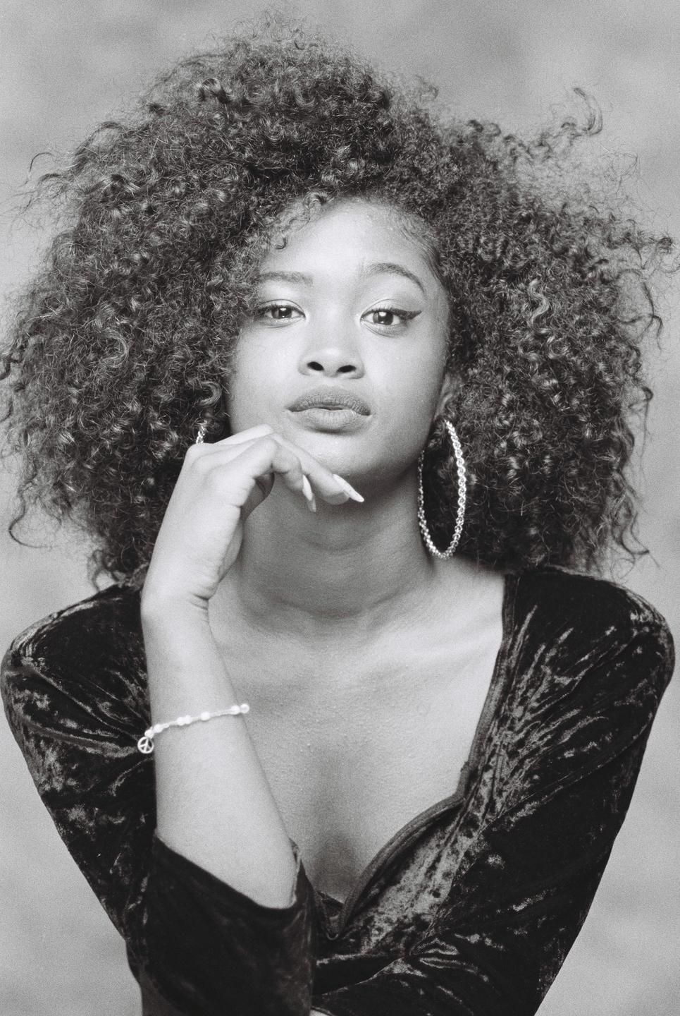 A woman with curly hair and hoop earrings poses with her hand on her chin.