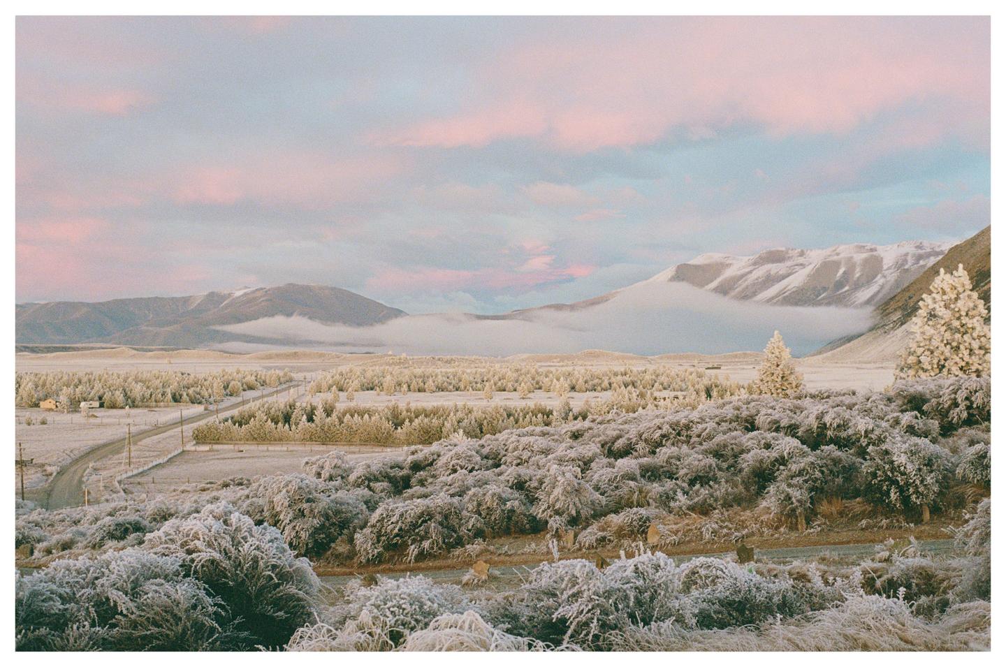 A winter landscape shows a road winding through a valley with snow-covered trees and mountains under a pink and blue sky.